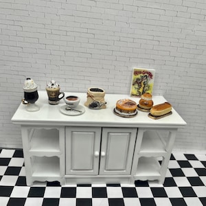 May include: Miniature coffee shop scene with a white table displaying various coffee drinks, pastries, and a bag of coffee beans. Includes a small sign with text. The scene is set against a white brick wall and a black and white checkered floor.