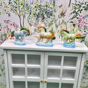 May include: Three ceramic horse figurines with rainbow manes and tails, standing on fluffy cloud bases. The horses are in shades of blue, tan, and green. They are displayed on a white cabinet with a floral wallpaper backdrop.