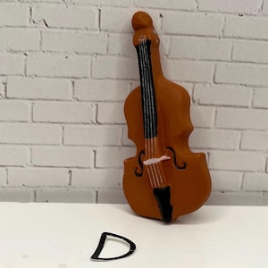 May include: A miniature brown double bass with black strings and a black tailpiece. The letter "D" is in front of the instrument. The background is a white brick wall.