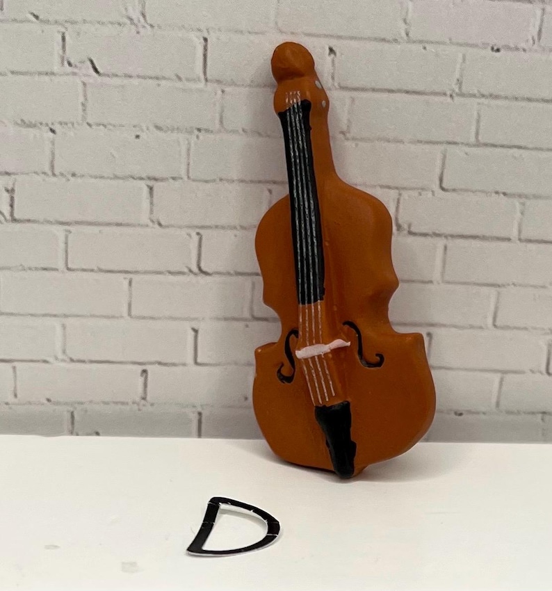 May include: A miniature brown double bass with black strings and a black tailpiece. The letter "D" is in front of the instrument. The background is a white brick wall.
