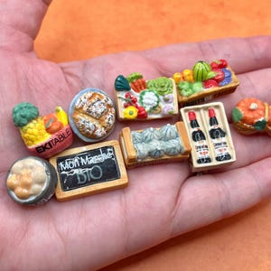 May include: Miniature ceramic food items including bread, produce, and wine bottles. A small chalkboard reads "Mon Marche BIO". The items are in a person's hand against an orange background. The items are likely for a dollhouse or display.