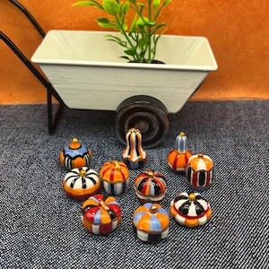 May include: A collection of miniature ceramic pumpkins in various colors and patterns, including orange, blue, and white. A white wheelbarrow with a green plant is in the background. The pumpkins are arranged on a denim surface.