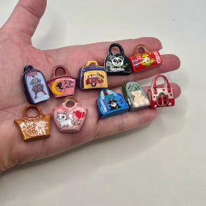 May include: A collection of miniature, colorful handbag charms. Each charm features a unique design, including a panda, a cat, a car, and a superhero. The charms are held in a hand, showcasing their small size and intricate details.