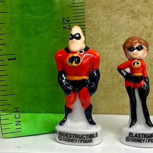 May include: Two small ceramic figurines of characters from The Incredibles. Mr. Incredible is in a red and black suit, and Elastigirl is in a similar outfit. The figurines are approximately 2 inches tall and are on white bases with character names and Disney/Pixar branding.