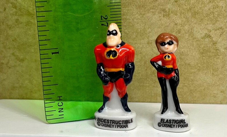 May include: Two small ceramic figurines of characters from The Incredibles. Mr. Incredible is in a red and black suit, and Elastigirl is in a similar outfit. The figurines are approximately 2 inches tall and are on white bases with character names and Disney/Pixar branding.