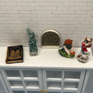 May include: Miniature ceramic figurines of Notre Dame de Paris, including a book, a statue, a stained glass window, a gnome, and a lion dancer. The figurines are displayed on a white cabinet against a white brick wall.