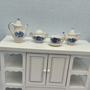 May include: Miniature white porcelain tea set with blue floral designs. The set includes a teapot, sugar bowl, teacup with saucer, and a creamer. The tea set is displayed on a white cabinet.