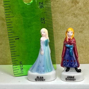 May include: Two small porcelain figurines of Elsa and Anna from Disney's Frozen. Elsa wears a light blue dress, and Anna wears a red cape over a blue dress. Each figurine is on a white base with the character's name and the Disney logo.