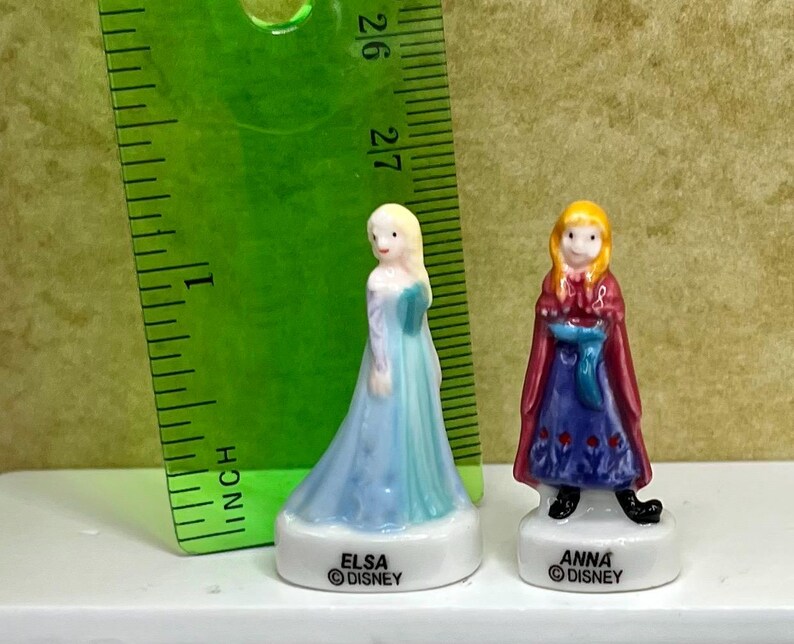 May include: Two small porcelain figurines of Elsa and Anna from Disney's Frozen. Elsa wears a light blue dress, and Anna wears a red cape over a blue dress. Each figurine is on a white base with the character's name and the Disney logo.