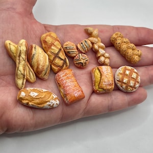 May include: A collection of miniature bread sculptures, including baguettes, loaves, and rolls, in various shapes and sizes. The bread pieces are painted with realistic colors, showcasing textures and details. The collection is held in a hand.