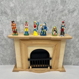 May include: A miniature wooden fireplace with a black grate and arched opening, topped with six small, colorful figurines. The figurines depict various characters, including a fairy, a wizard, and a soldier. The fireplace is made of light-colored wood.