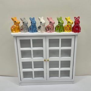 May include: A white cabinet with glass-paneled doors, topped with seven colorful ceramic bunny figurines. The bunnies are in various colors, including yellow, gray, blue, white, pink, green, and red. The cabinet has gold-colored door knobs.