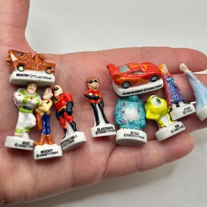 May include: A collection of small, colorful Disney and Pixar character figurines, including Buzz Lightyear, Woody, and characters from The Incredibles and Cars. Each figurine is mounted on a white base with the character's name.