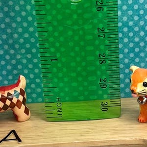 May include: Two small, decorative ceramic figurines of animals. One is a dog with a red and brown checkered pattern, and the other is a cat with orange and yellow floral designs. A green ruler is placed between them, measuring approximately 3 inches.