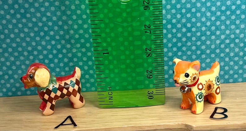 May include: Two small, decorative ceramic figurines of animals. One is a dog with a red and brown checkered pattern, and the other is a cat with orange and yellow floral designs. A green ruler is placed between them, measuring approximately 3 inches.