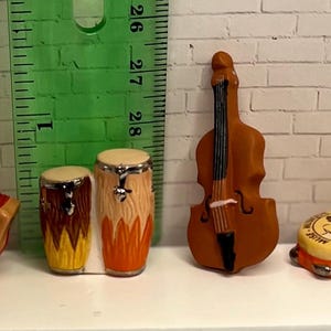 May include: Miniature musical instruments including a red accordion, two drums, a brown upright bass, and a tambourine. The tambourine has a yellow top with a black and white design and silver jingles. The instruments are on a white surface.