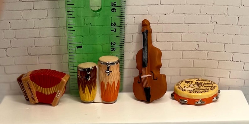 May include: Miniature musical instruments including a red accordion, two drums, a brown upright bass, and a tambourine. The tambourine has a yellow top with a black and white design and silver jingles. The instruments are on a white surface.