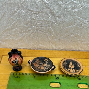 May include: Miniature ceramic Greek pottery pieces. The set includes a small vase, a bowl with handles, and a round plate. Each piece features black and orange designs depicting figures and scenes from Greek mythology. The vase is approximately 1 inch tall.