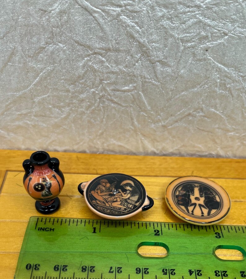May include: Miniature ceramic Greek pottery pieces. The set includes a small vase, a bowl with handles, and a round plate. Each piece features black and orange designs depicting figures and scenes from Greek mythology. The vase is approximately 1 inch tall.