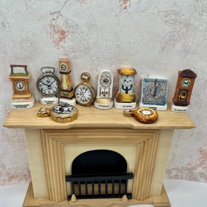 May include: A collection of miniature ceramic clock figurines displayed on a wooden mantelpiece. The set includes various styles such as grandfather, carriage, and pocket watches, each with printed labels like La Pendulette de Voyage and Le Carillon.