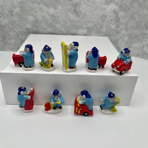 May include: A collection of miniature ceramic figurines depicting firefighters in blue uniforms. The figurines are engaged in various activities, including driving a red fire truck, using a ladder, and working with a fire hydrant. The figurines are on a white surface.
