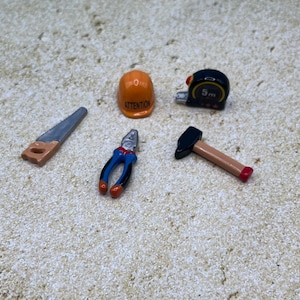 May include: Miniature construction tools including a saw, pliers, hammer, tape measure, and an orange hard hat with the word "ATTENTION". The tape measure reads 5m. The tools are arranged on a textured surface.
