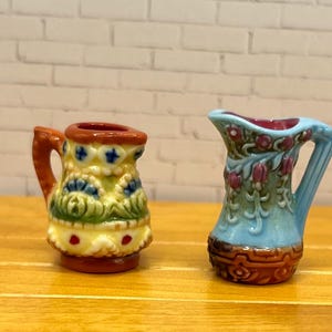 May include: Two miniature ceramic pitchers. One is yellow, green, and orange with a floral design. The other is light blue with a floral pattern and a brown base. Both have handles and are on a wooden surface.