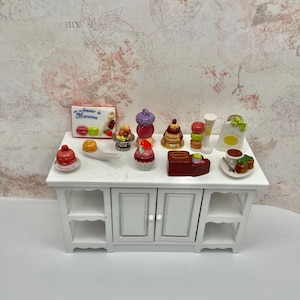 May include: A white miniature kitchen cabinet displays various miniature desserts, including macarons, cupcakes, and chocolates. A sign reads "Amour & Macarons." The desserts are in shades of red, purple, yellow, and brown. The cabinet has two doors and open shelves.