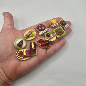 May include: A collection of miniature, colorful dessert figurines, including cakes, pastries, and chocolates. Some pieces have text, such as "MERCHANT AU CHOCOLAT" and "AU CHOCOLAT". The desserts are arranged on a light background.