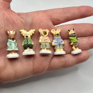 May include: Five miniature animal figurines, including a rabbit, deer, and three mice, each dressed in detailed clothing. The figurines are painted with bright colors and stand on small white bases. The collection is held in a hand.