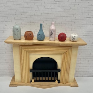 May include: A miniature wooden fireplace mantel with a light wood finish, supporting six small decorative ceramic vases. The vases are in various colors, including blue, pink, and red, and feature floral and face designs in black.