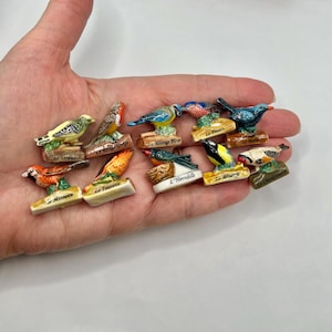 May include: A collection of ten miniature ceramic bird figurines, each perched on a small base labeled with French names including Le Verdier, Le Rouge-Gorge, La Mesange Bleue, Le Pinson, Le Merle, Le Moineau, La Fauvette, L'Hirondelle, La Mesange, and Le Chardonneret.