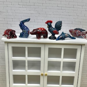 May include: A collection of colorful, hand-painted animal figurines, including a turtle, rabbit, bear, rooster, dolphin, and fish, displayed on a white cabinet. The figurines feature intricate patterns and vibrant colors, adding a decorative touch.