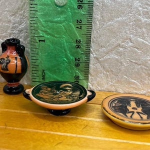 May include: Miniature ceramic Greek pottery pieces, including a vase and two plates. The vase is black and orange, and the plates feature detailed scenes. A green ruler is in the background, showing measurements in inches.