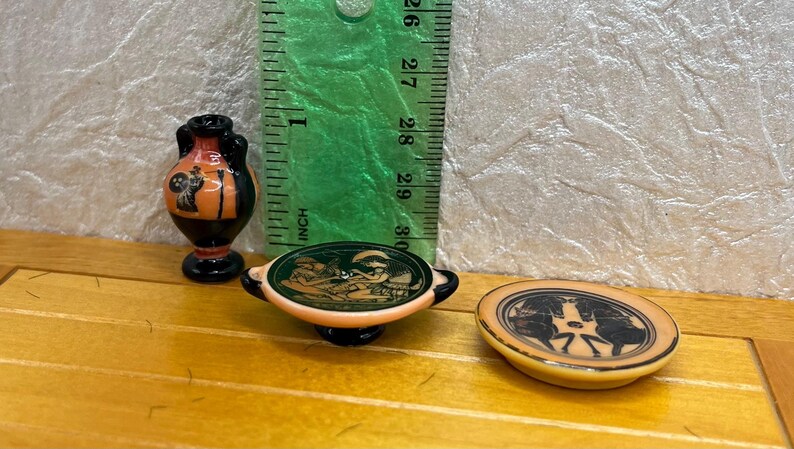 May include: Miniature ceramic Greek pottery pieces, including a vase and two plates. The vase is black and orange, and the plates feature detailed scenes. A green ruler is in the background, showing measurements in inches.