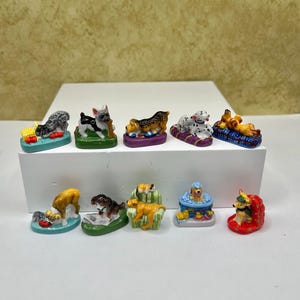 May include: A collection of small, colorful dog figurines. Each figurine depicts a different breed of dog in various poses, such as lying down or sitting. The figurines are displayed on a white surface, with a neutral background.