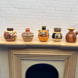 May include: Five small, decorative ceramic vases with hand-painted designs. The vases are in shades of orange, brown, and black, with geometric and figurative patterns. They are displayed on a light-colored wooden surface.