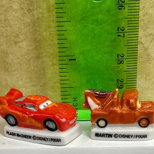 May include: Two miniature toy cars, Flash McQueen and Mater, from the Disney Pixar Cars franchise. Flash McQueen is red with yellow flames, and Mater is brown. Both are on white bases with their names.