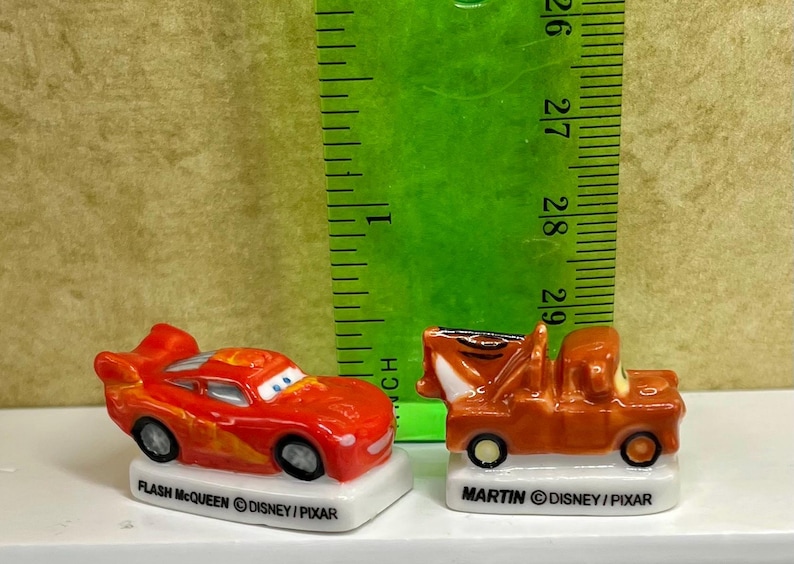 May include: Two miniature toy cars, Flash McQueen and Mater, from the Disney Pixar Cars franchise. Flash McQueen is red with yellow flames, and Mater is brown. Both are on white bases with their names.