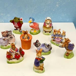 May include: A collection of ceramic figurines featuring various animal characters. The figurines include a frog, duck, rabbit, cats, and hedgehogs, all in colorful outfits. The figurines are arranged on a white surface.