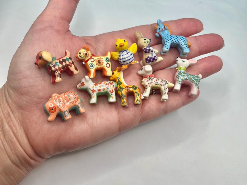 May include: A collection of miniature animal figurines, including dogs, a giraffe, a bunny, a deer, and a donkey. Each figurine is brightly colored with intricate patterns and designs, held in a person's hand.