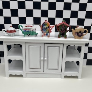 May include: A white cabinet with two doors and open shelves, displaying five miniature decorative teapots. The teapots are various colors and designs, including a red and white teapot, a green and white teapot, a colorful teapot, a cuckoo clock teapot, and a tan teapot.