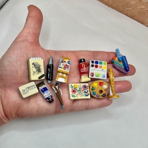 May include: Miniature artist's supplies including a sketchbook, paint tubes, brushes, a palette, and a small easel. The set includes a small statue and a block of paint. The items are in various colors.