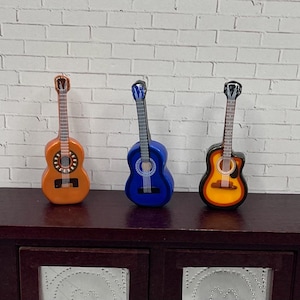 May include: Three miniature acoustic guitars in orange, blue, and yellow-brown. The guitars are displayed on a dark brown wooden surface against a white brick wall. Each guitar has a detailed design, including strings and sound holes.