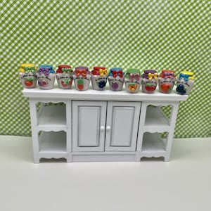 May include: A white miniature cabinet with a shelf and two doors, topped with nine small ceramic jars. Each jar has a different fruit design and a colorful bow on the lid. The jars are arranged in a row.