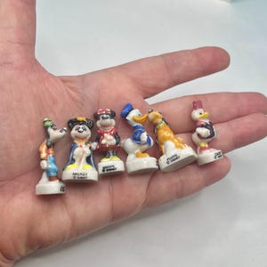 May include: A collection of six small Disney character figurines, including Goofy, Mickey Mouse, Minnie Mouse, Donald Duck, Pluto, and Daisy Duck. Each figurine is painted with bright colors and stands on a white base.