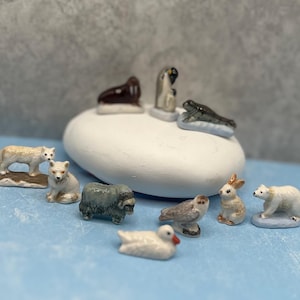 May include: A collection of miniature ceramic animal figurines, including a wolf, fox, musk ox, owl, rabbit, polar bear, duck, penguin, seal, and frog. The figurines are painted in realistic colors and set on a blue surface.