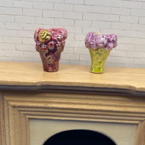 May include: Two miniature ceramic vases. One is a deep pink with gold accents, the other is light purple with a green base. Both are on a wooden surface against a white brick wall. The vases have a textured, floral design.