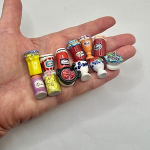 May include: A collection of miniature ceramic food items including small jars with labels like Mûre, Cerises, and Miel, a black pot with red contents, white bowls with blue floral patterns, and a small dish with blue and green fruit decorations.