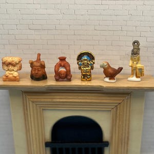 May include: A collection of miniature ceramic figurines, including a bird, a deity, and other ancient-style sculptures, displayed on a wooden shelf. The figurines are in shades of brown, gold, and white, with intricate details.